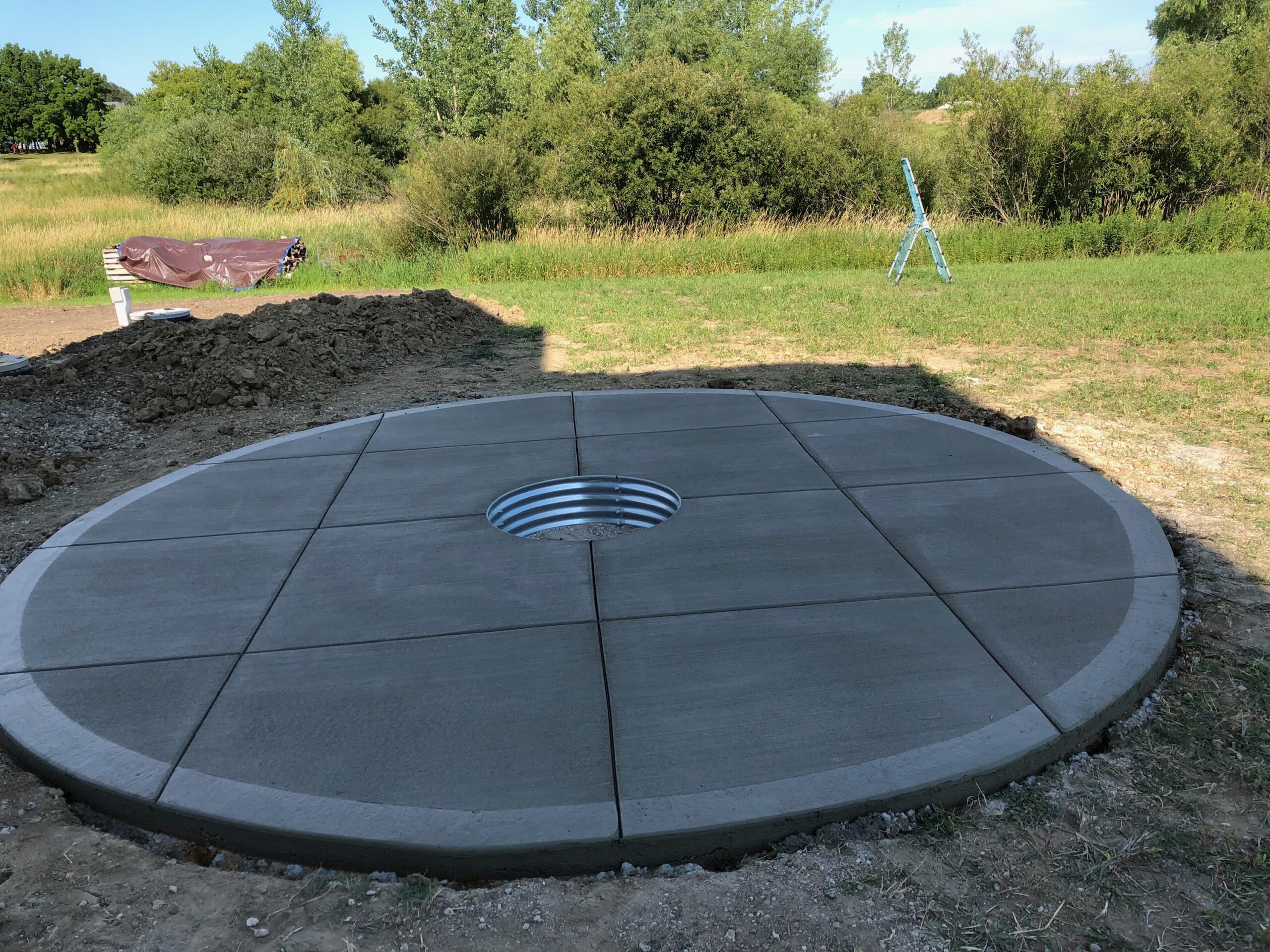 Concrete slab with a circular design and a central drainage hole, surrounded by unlandscaped earth, indicating groundwork for a construction project.