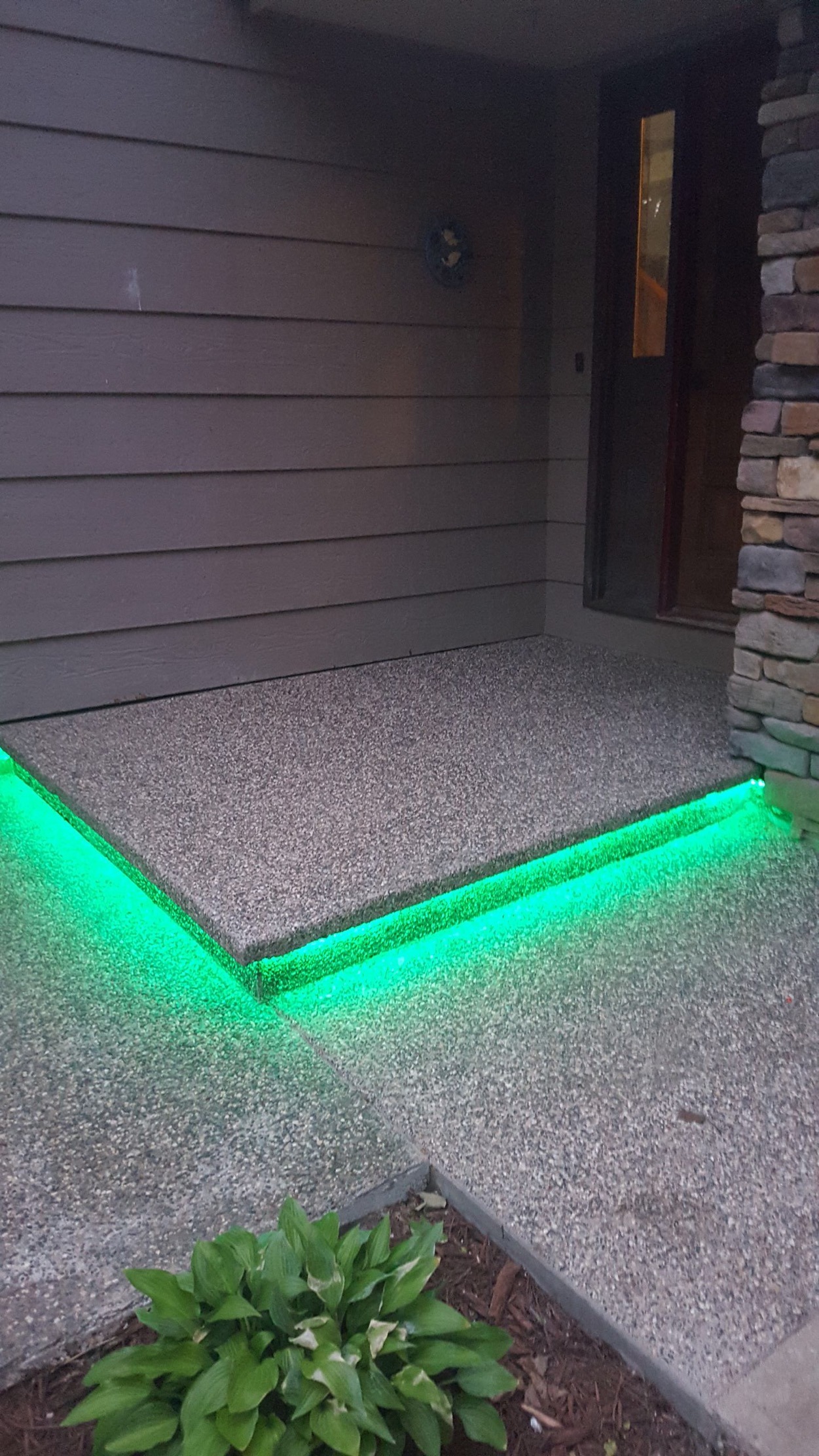 A well-lit exterior entrance features a textured concrete porch with vibrant green LED lighting along the edges, enhancing its modern aesthetic.