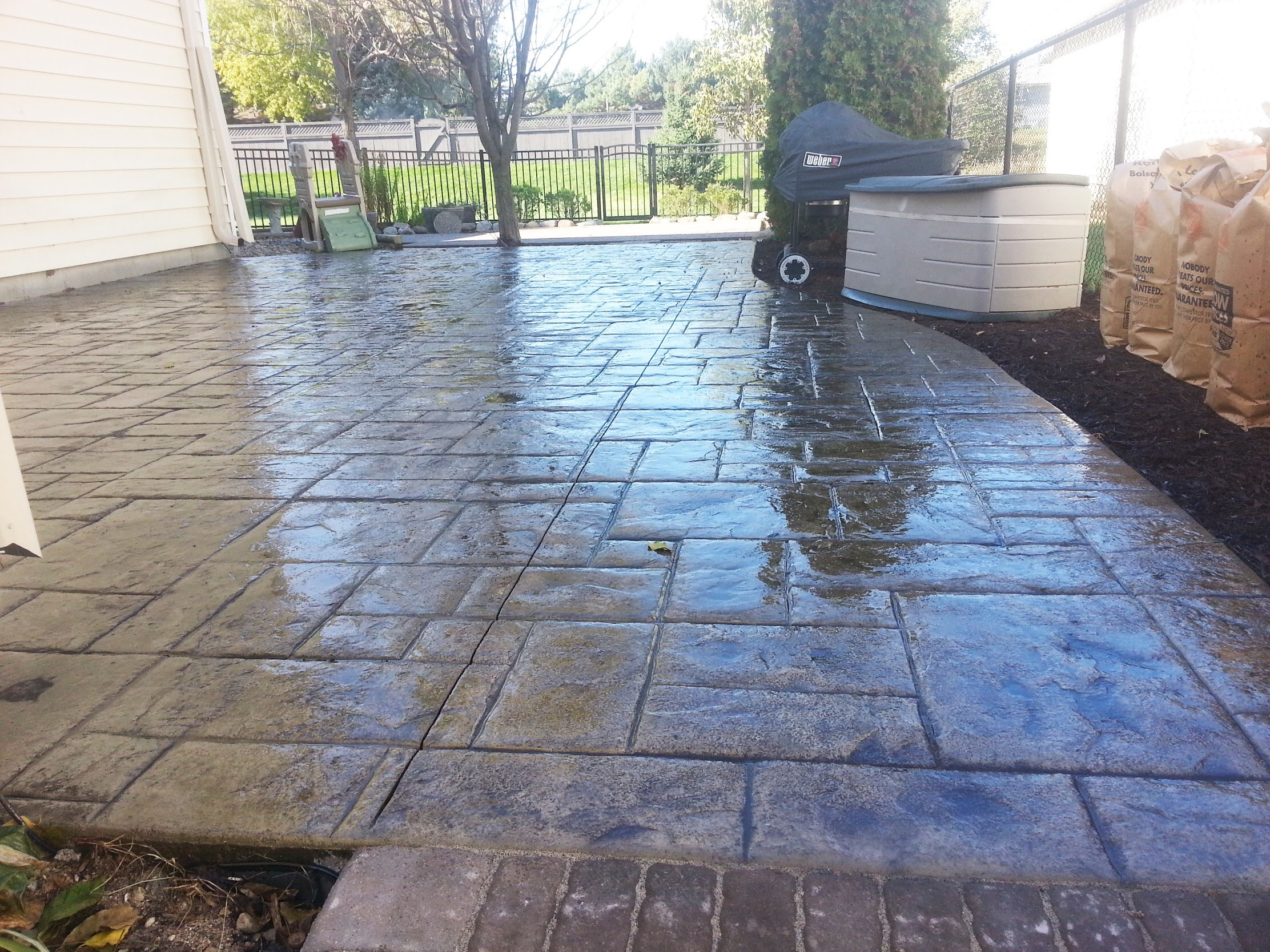 Textured, wet concrete patio featuring a grill and storage unit, surrounded by a well-maintained yard and fencing. Ideal for outdoor gatherings.
