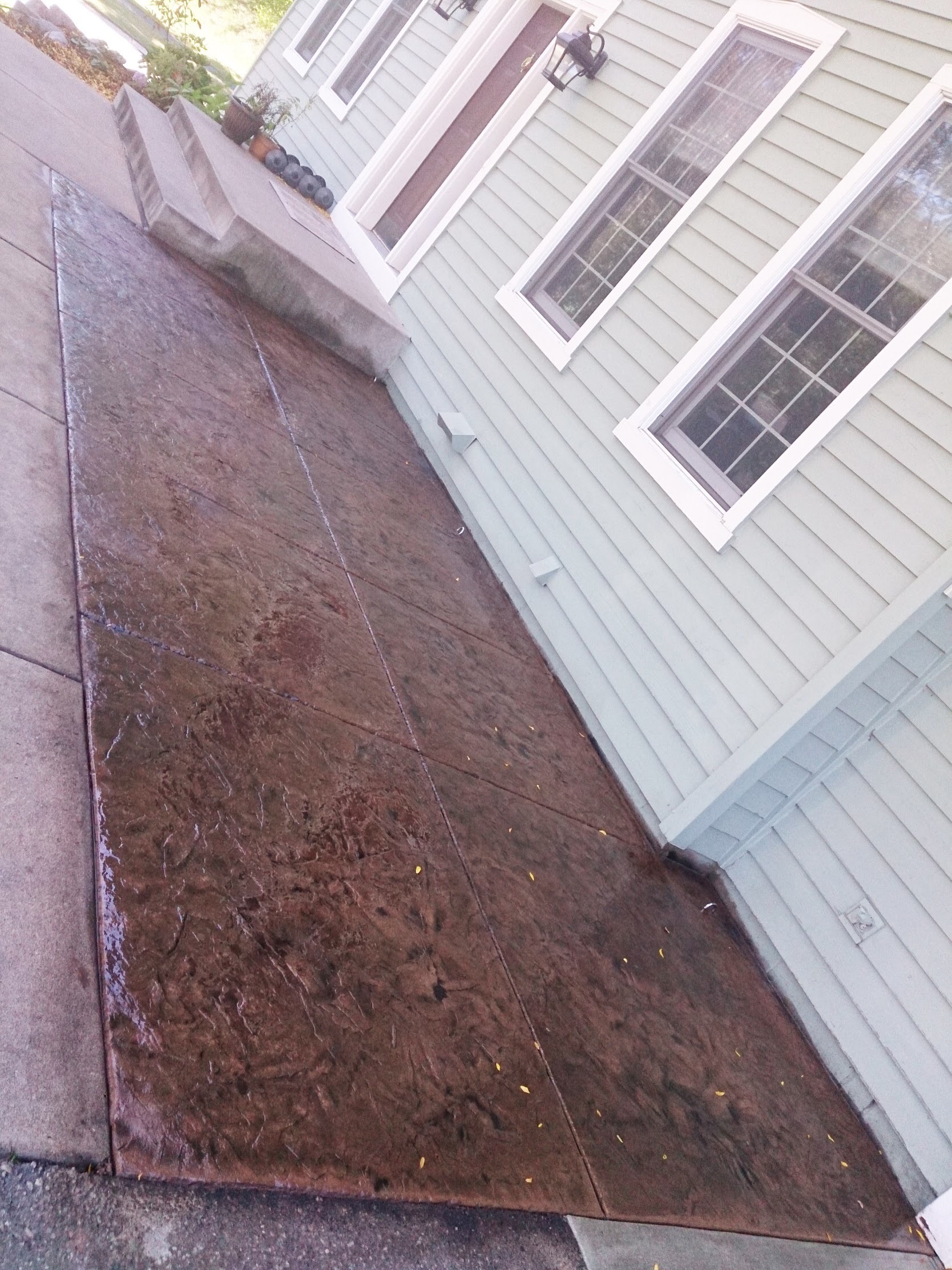 Newly poured and stamped concrete walkway leading to a home, featuring textured brown surface, adjacent to steps and a light green house.
