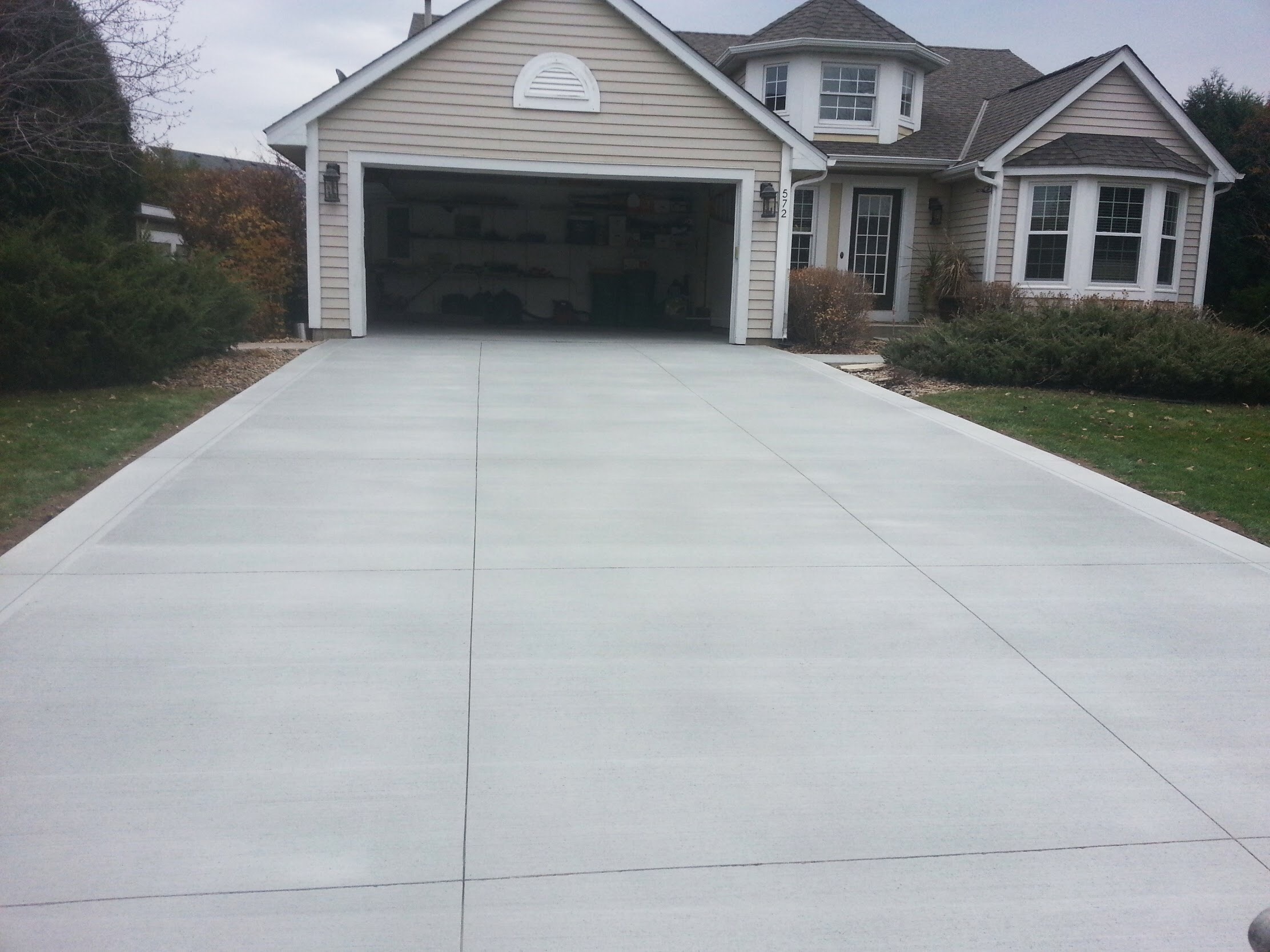 Freshly poured concrete driveway leading to a garage, framed by a well-maintained lawn and bushes, showcasing a home exterior.