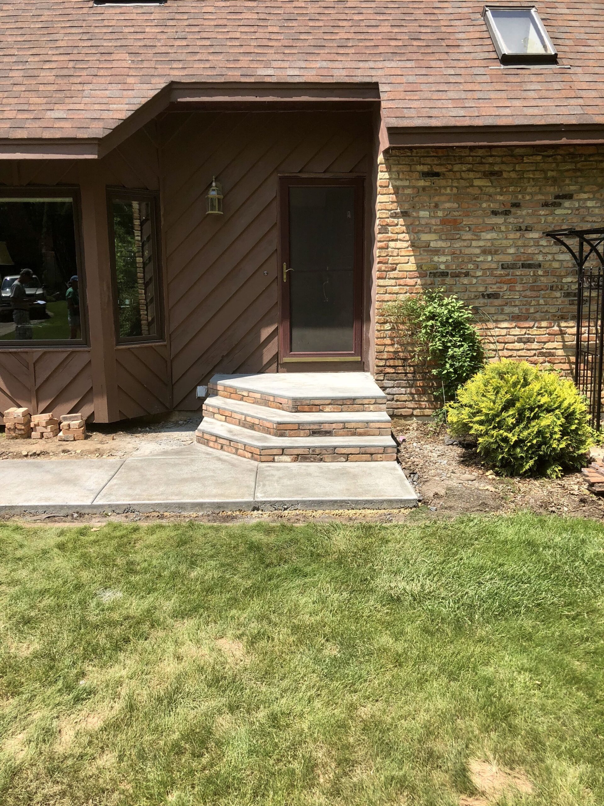 Newly constructed brick steps lead to a glass door entrance, framed by brown siding and manicured landscaping. The image highlights home accessibility.