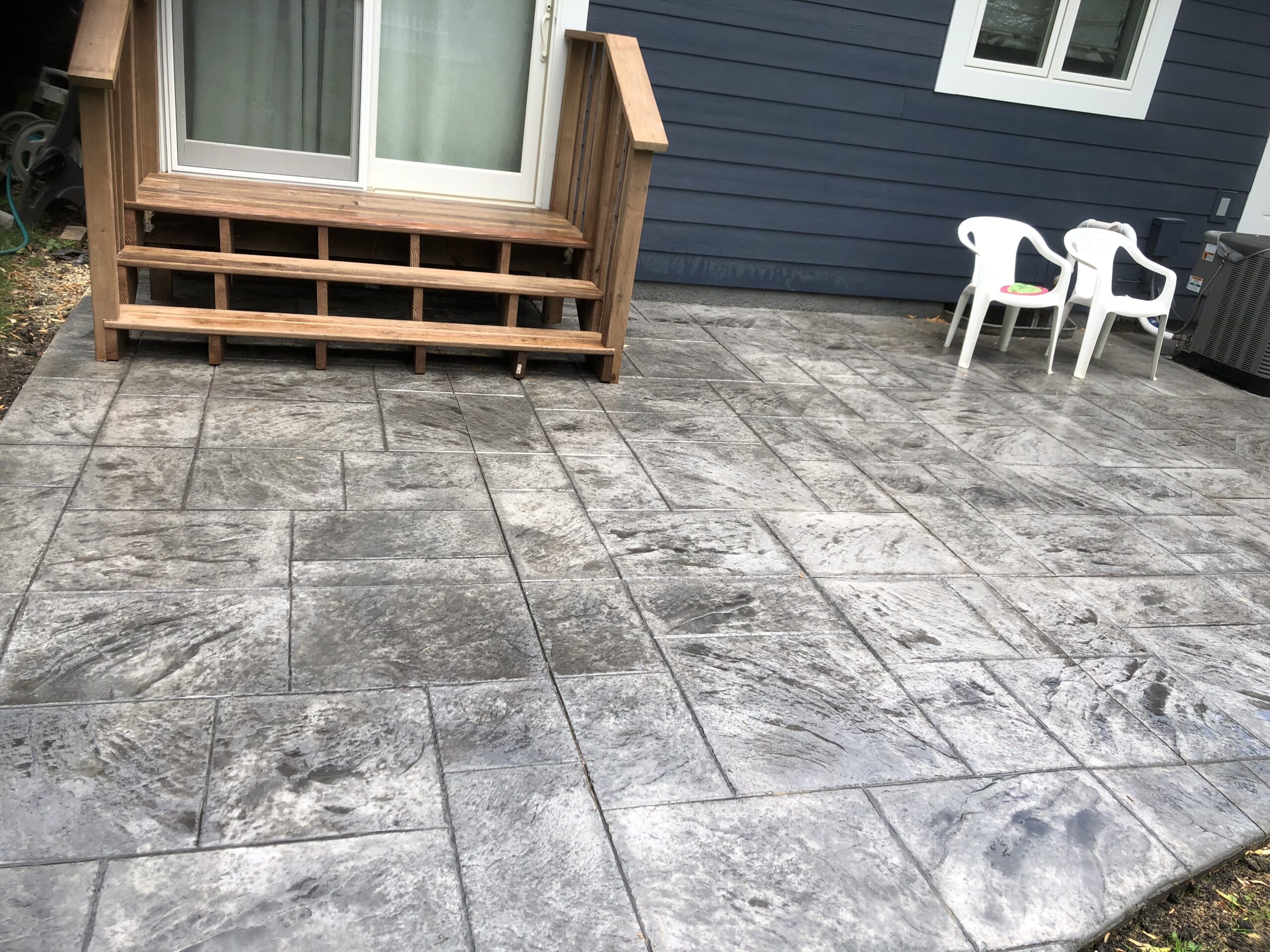 Textured gray stamped concrete patio with steps leading to a door, flanked by two white chairs, offering an outdoor relaxation space.