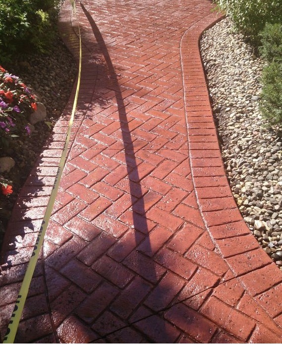 Curved, red brick pathway with a herringbone pattern, bordered by flowers and gravel. Damp surface reflects sunlight, enhancing its appeal.