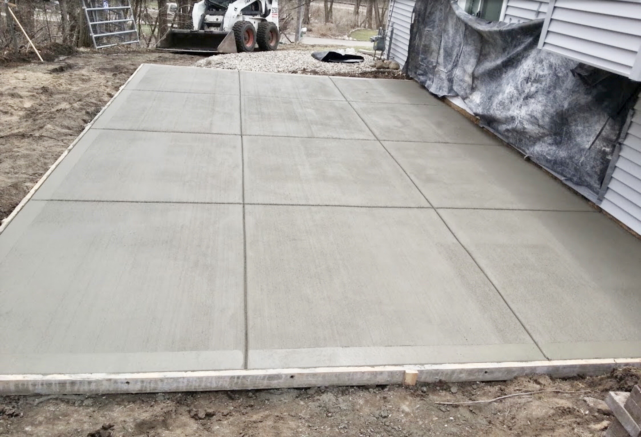 Freshly poured concrete slab, marked with lines for joints, in a residential outdoor space. Equipment in the background indicates ongoing construction work.