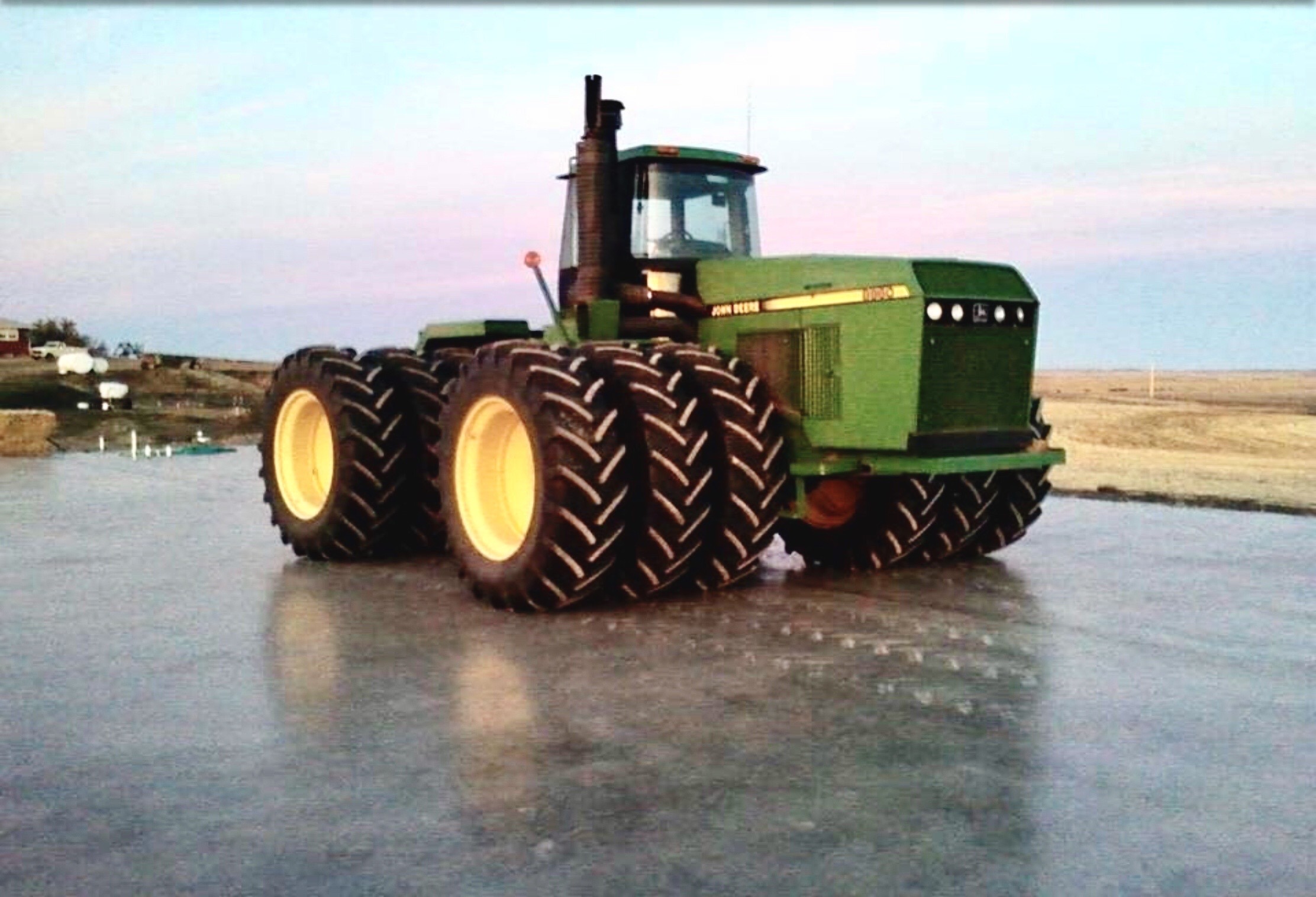 John Deere tractor with large tires parked on a concrete surface, set against a rural landscape at dusk. Highlights farming equipment versatility.
