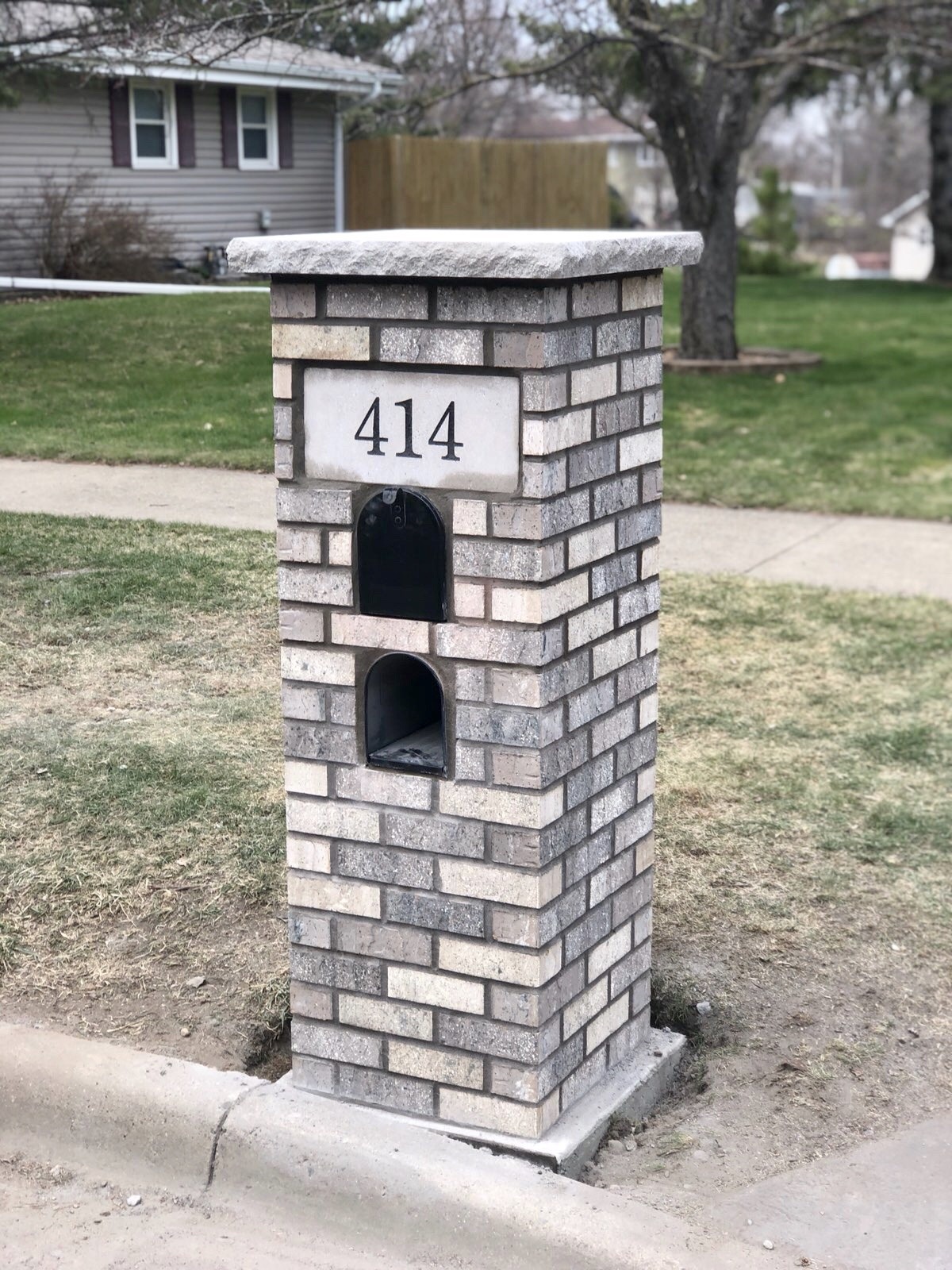 Brick mail pillar labeled "414" with a small mailbox opening, situated near a residential yard and sidewalk. Represents a distinctive home address marker.