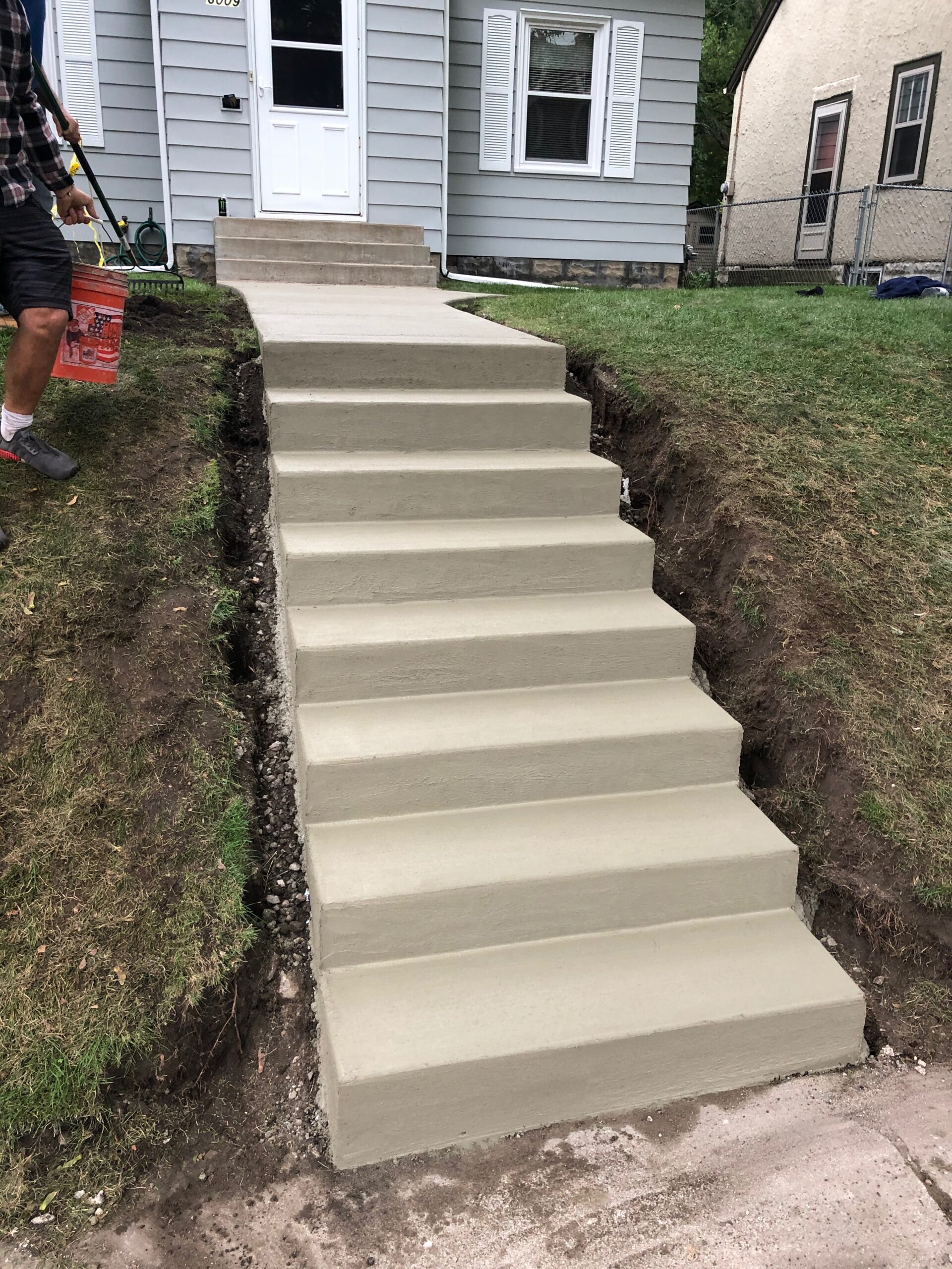 Newly constructed concrete steps lead to a house entrance, with a person holding a paint bucket nearby. The surrounding area is freshly graded.