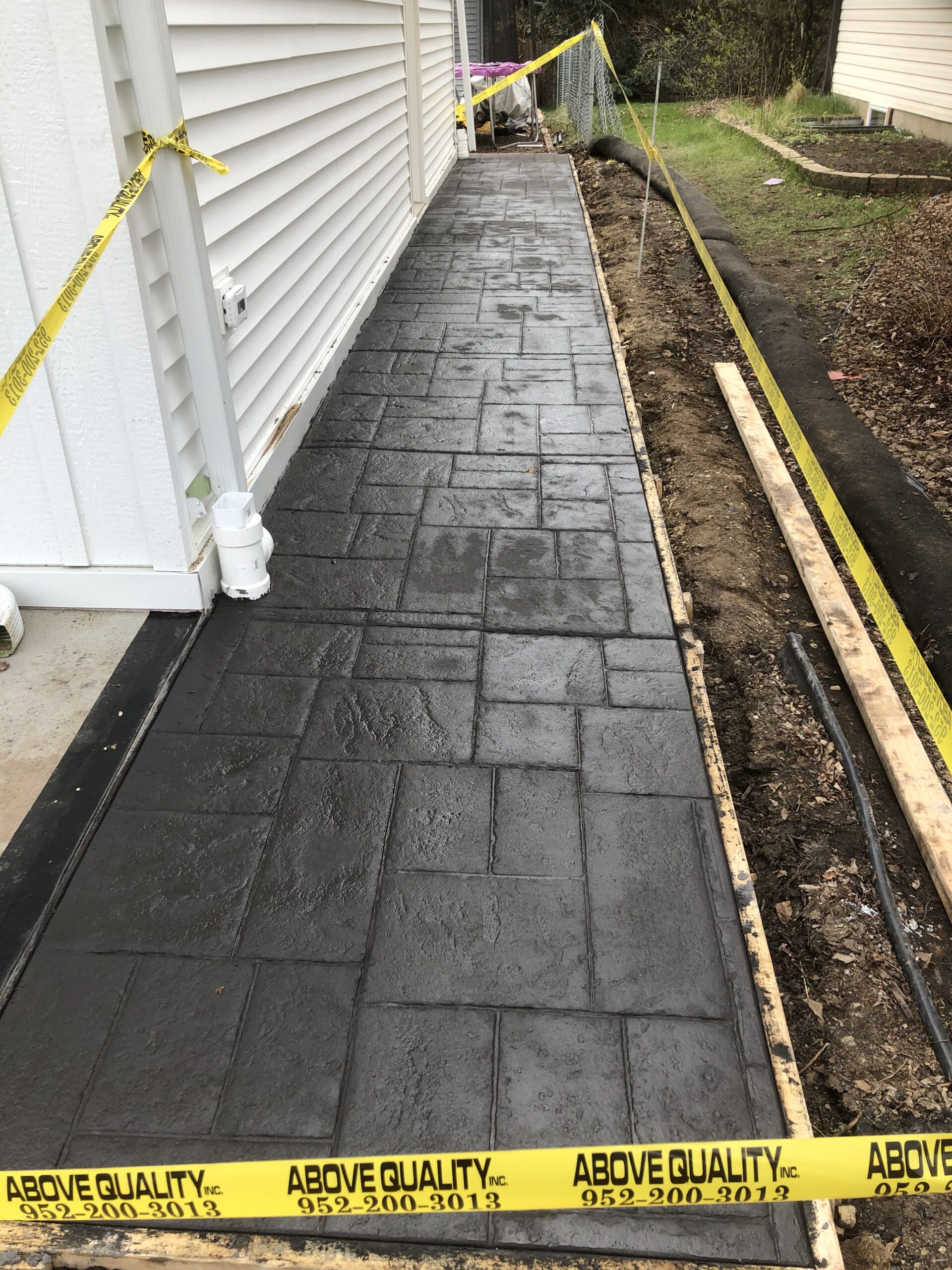 Freshly poured concrete sidewalk with a textured pattern, bordered by caution tape. Nearby, construction materials hint at ongoing yard work.